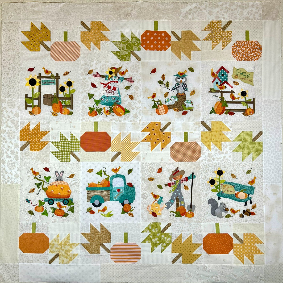 A square quilted fabric piece featuring a fall harvest theme with stitched pumpkins, leaves, scarecrows, a birdhouse, a tractor, and woodland animals on an off-white patterned background.